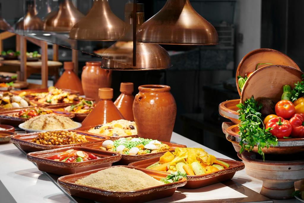 Brokastu bufete ar dažādiem ēdieniem, ieskaitot dārzeņu salātus, graudus un gaļu, pasniegta tradicionālajos traukos. Apgaismojums rada siltu un viesmīlīgu atmosfēru.