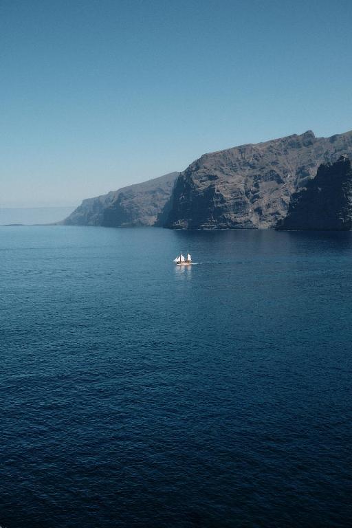 Kāds kuģis, ar trim burām, peld pa mierīgu jūru, apkārt augstām klintīm un skaidrām debesīm.