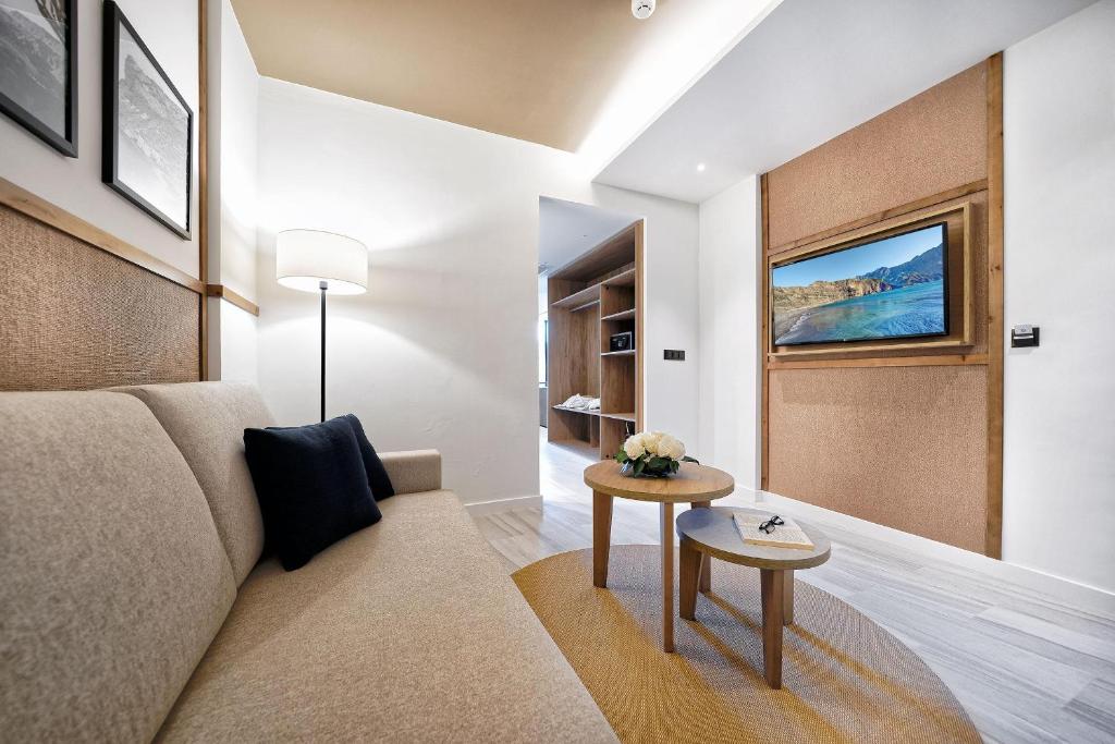 Mūsdienīgs viesistabas interjers ar gaišām krāsām, dīvānu un televizoru uz sienas, kas papildina telpas komfortu. Uz galdiņa atrodas ziedi un brilles, radot mājīgu atmosfēru.