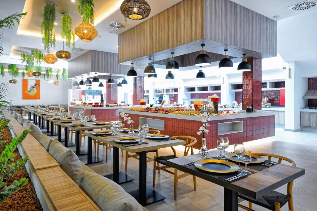 Mūsdienīgs restorāns ar elegantiem galdiem un siltu apgaismojumu, kas rada patīkamu atmosfēru. Uz galda ir izkārtoti trauki un ziedi, bet fona redzama bagātīga ēdienu izvēle.