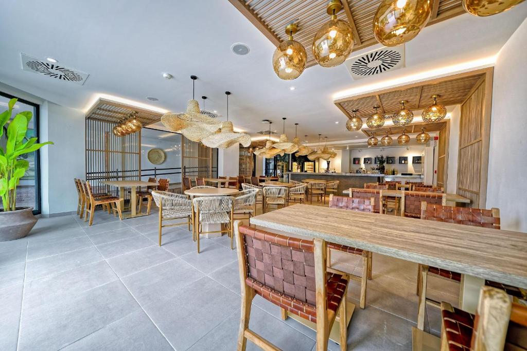 Mūsdienīgs restorāna interjers ar koka mēbelēm un stilīgām lampām, kas radījušas siltu atmosfēru. Gaisma un zaļa auga klātbūtne piešķir telpai dzīvīgumu.