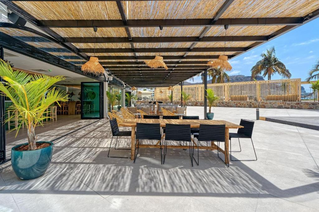 Āra terase ar koka galdiem un krēsliem, rotāta ar palmu lapām un dabīgām dekorācijām, nodrošina relaksējošu atmosfēru. Pārklājums nodrošina ēnu, kamēr fonā redzamas palmas un skaidras debesis.