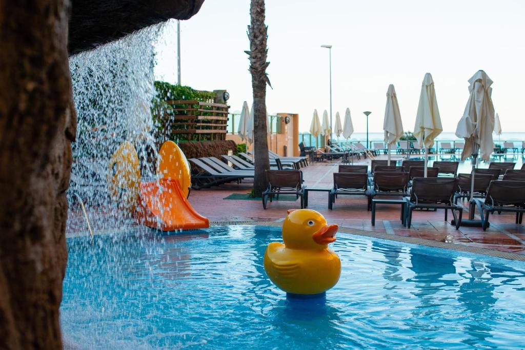 Dzeltenā plastmasas pīle peld baseinā, kas atrodas viesnīcas teritorijā ar ūdens kritumu un slidkalniņu. Fons satur saulessargus un sauļošanās krēslus.