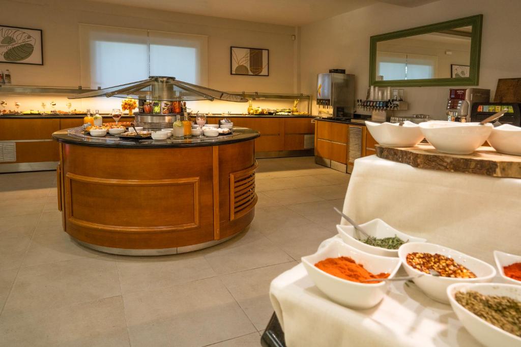 Mūsdienīgs bufetes stils ar dažādām garšvielām un dzērieniem, novietots centrālā apaļā galda. Interjers ar koka akcentiem un gaišām krāsām, radot viesmīlīgu atmosfēru.