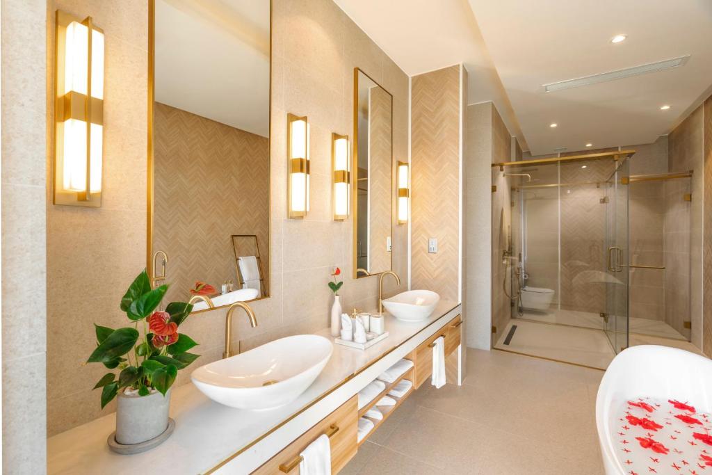 Mūsdienīgs vannas istabas interjers ar elegantiem zelta aksesuāriem, divām izlietnēm un stikla dušu. Telpā ir dabīgas dekorācijas un silts apgaismojums, radot relaksējošu atmosfēru.