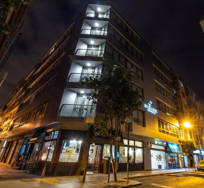 Mūsdienīga ēka ar apgaismotiem balkoniem, kas atrodas pilsētas ielā naktī. Zemāk redzami kafejnīca un citas komerciālās telpas.