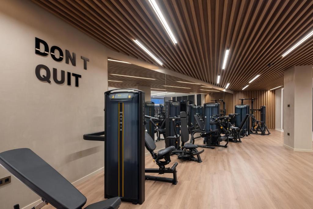 Mūsdienīgs sporta zāles interjers ar sporta aprīkojumu un motivējošu uzrakstu "DON'T QUIT" uz sienas. Gaismas un koka elementu kombinācija rada dinamisku un iedvesmojošu vidi treniņiem.