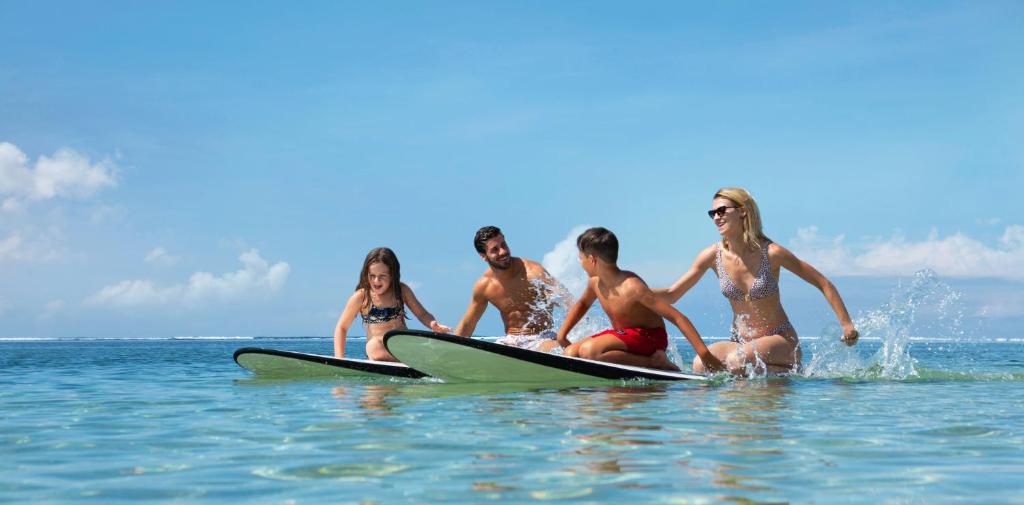 Četri cilvēki, divi pieaugušie un divi bērni, izbauda sērfošanu mierīgā jūrā ar saulainām debesīm. Viņi smejas un rotaļājas uz sērfošanas dēļiem, radot priecīgu atmosfēru.