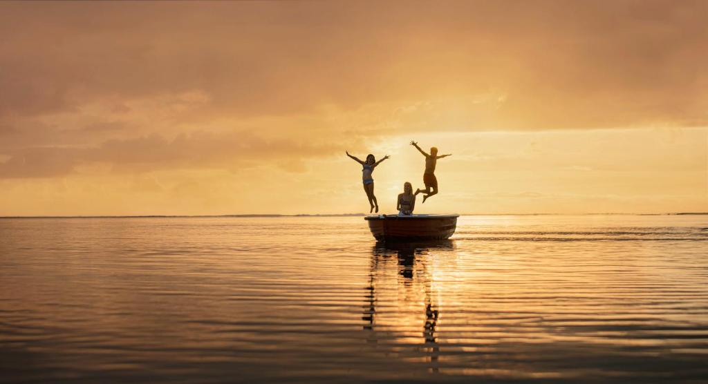 Trīs cilvēki priecīgi lēkā laivā saulrieta gaismā, atspoguļojoties mierīgajā ūdenī.