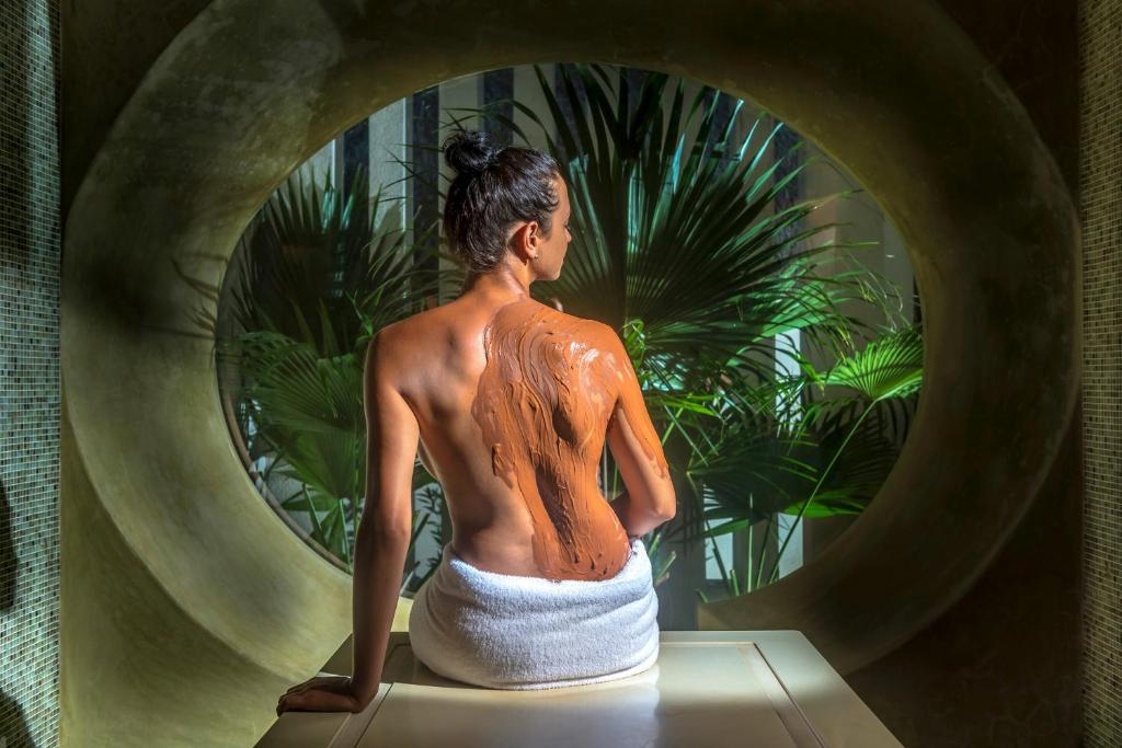 Sieviete ar ķermeņa masku sēž relaksējošā spa vidē, kamēr fons ir rotāts ar zaļām palmu lapām.