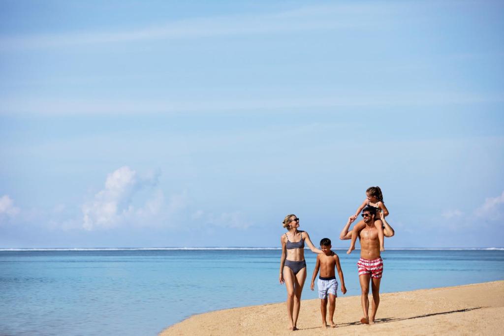 Ģimene pastaigā pa pludmali, baudot saulainu dienu. Fons veido mierīgs jūras skats ar ziliem debesīm un mīkstiem mākoņiem.