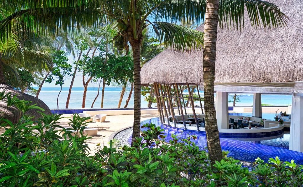 Tropu kūrorta ainava ar bāru, kas atrodas pie pludmales, ieskauta palmām un ziediem, radot relaksējošu atmosfēru. Skats uz jūru un saulainu debesi papildina mierīgu brīvdienu sajūtu.