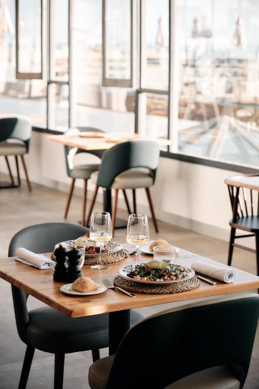 Elegants restorāna interjers ar koka galdu, uz kura ir pasniegts ēdiens, divi vīna glāzes un saldums. Logu aizmugurē redzams pilsētas skats, radot patīkamu atmosfēru.