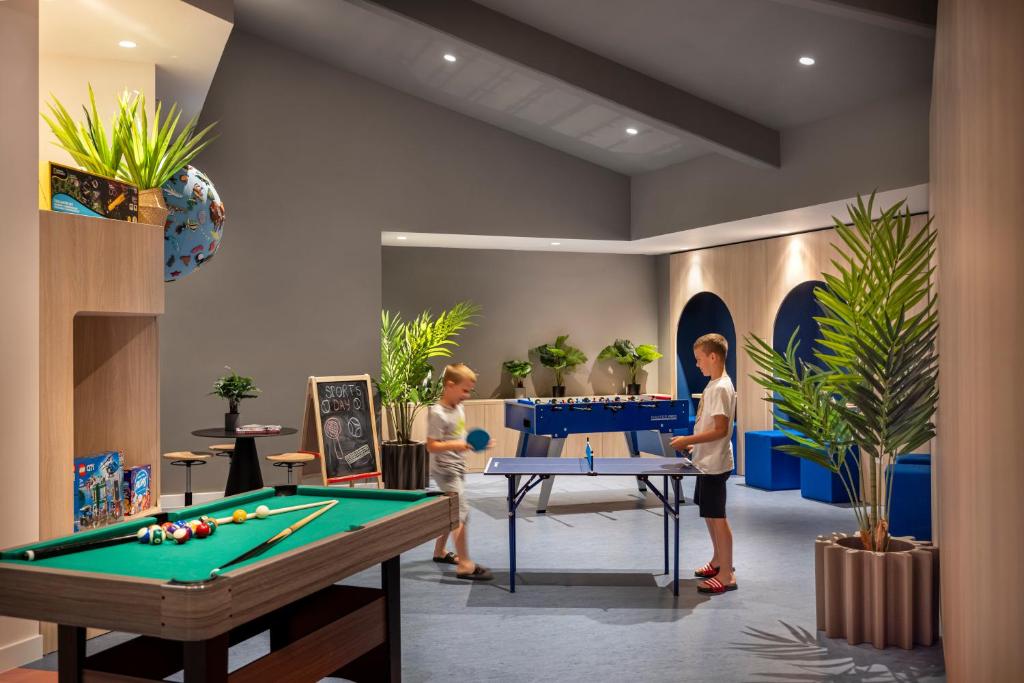 Mūsdienīga spēļu telpa ar biljarda galdu un galda tenisa galdu, kur divi bērni spēlē un izbauda brīvo laiku. Telpā ir arī zaļas augu dekorācijas un mūsdienīgs interjers.
