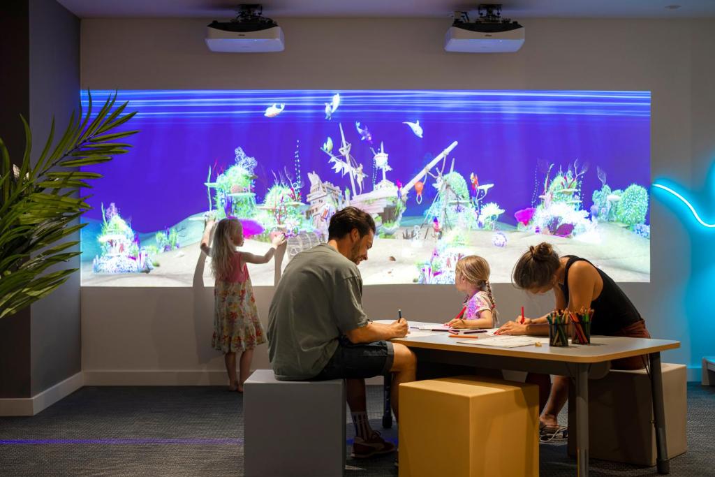 Bērni un pieaugušie kopā radoši darbojas pie galda, zīmējot, kamēr interaktīva projekcija rāda krāsainu jūras ainavu.