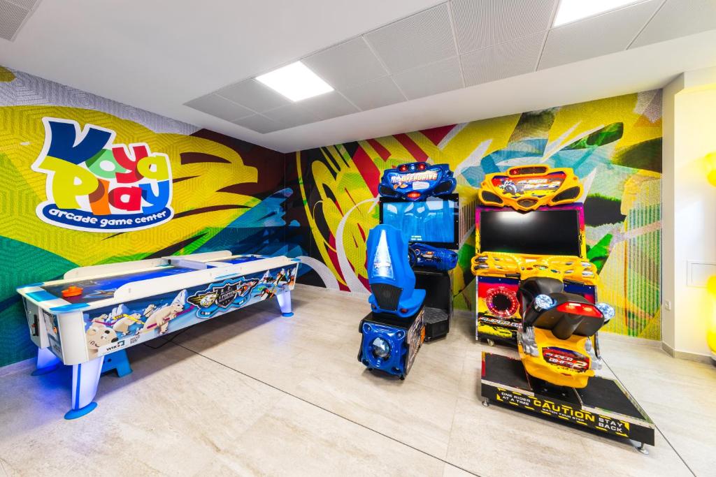 Krāsains spēļu centrs ar Vaya Play logo, kurā redzamas dažādas arcade spēles, tostarp gaisa hokeja galda un motocikla spēļu automāti.