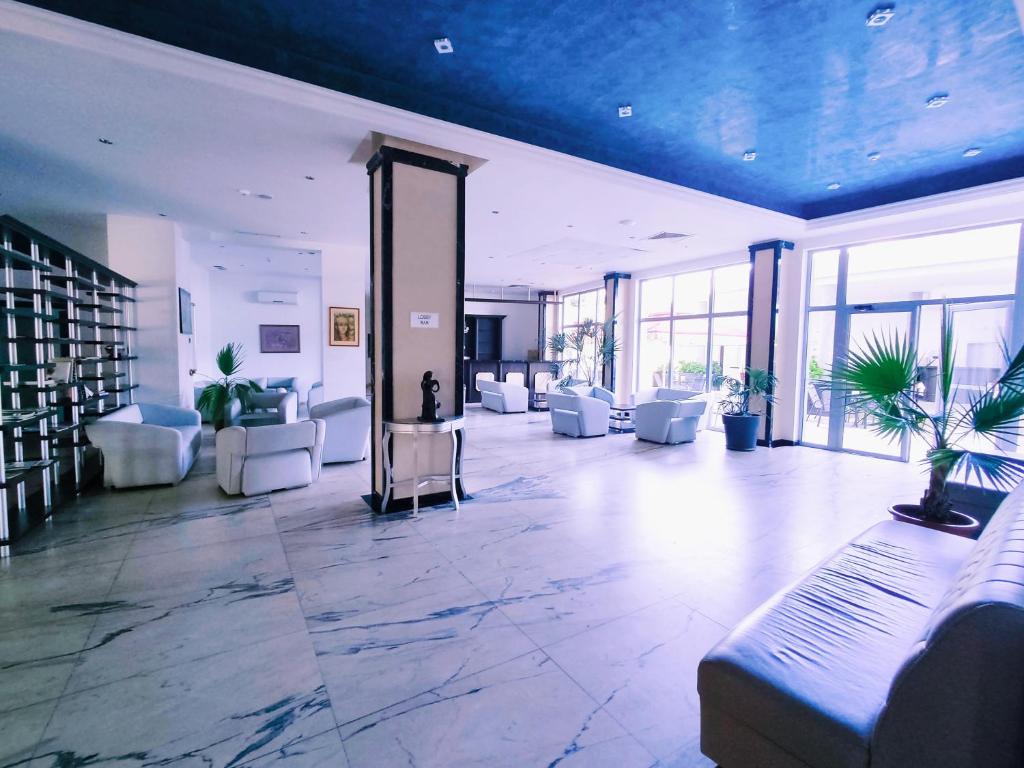 Mūsdienīgs viesnīcas vestibils ar ērtām baltām mēbelēm un lieliem logiem, kas nodrošina dabisko apgaismojumu. Elegantais dizains un zaļās augu dekorācijas rada relaksējošu atmosfēru.