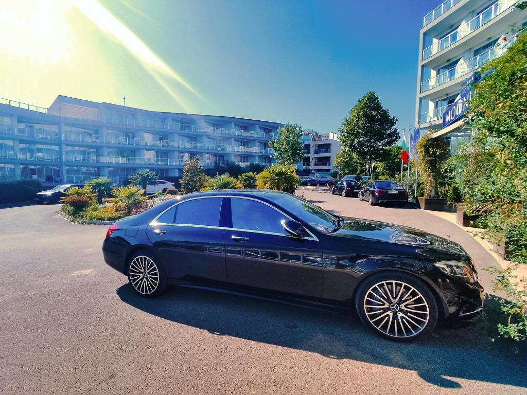 Melns luksusa auto stāv pie viesnīcas, iepriekšējā fona ir moderns arhitektūras komplekss ar zaļām ainavām un saules gaismu.