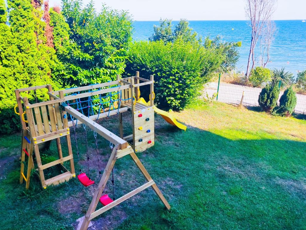 Bērnu rotaļu laukums ar šūpolēm, slidkalniņu un kāpšanas sienu, izvietots dārzā pie jūras. Apkārt aug zaļi krūmi un koki, radot patīkamu atmosfēru.
