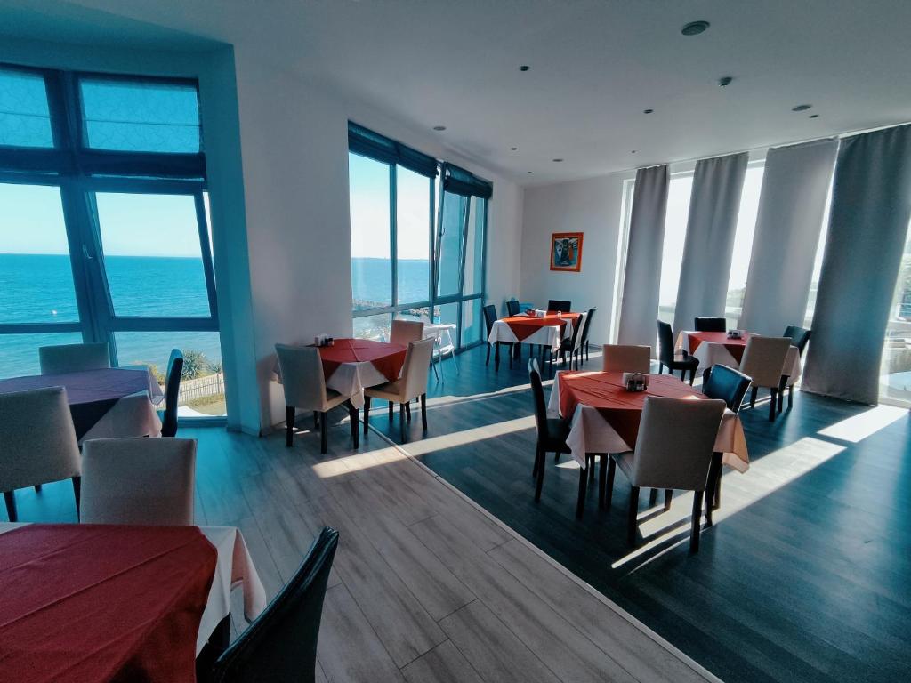 Gaiša restorāna telpa ar skatu uz jūru, kur ir ēdamgaldi ar sarkaniem un baltiem galdautiem. Labi izgaismota vide ar lielām logu sienām un mūsdienīgu interjeru.