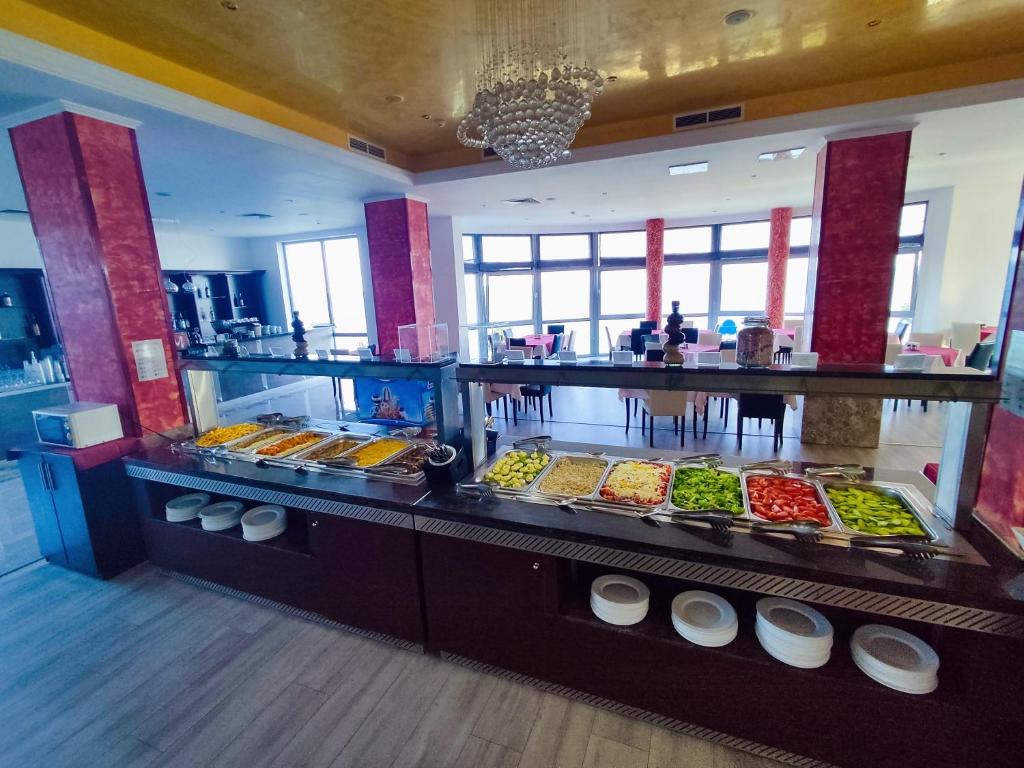 Bufete ar dažādiem ēdieniem un salātiem, redzami skaisti dekorēti galdi un lieli logi ar skatu uz ārpusi.