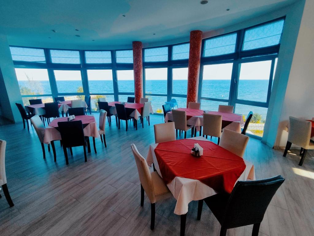 Restorāna telpa ar jūras skatu, kurā ir galdi ar sarkaniem un baltiem galdautiem. Gaisma plūst caur lielajiem logiem, radot patīkamu atmosfēru.