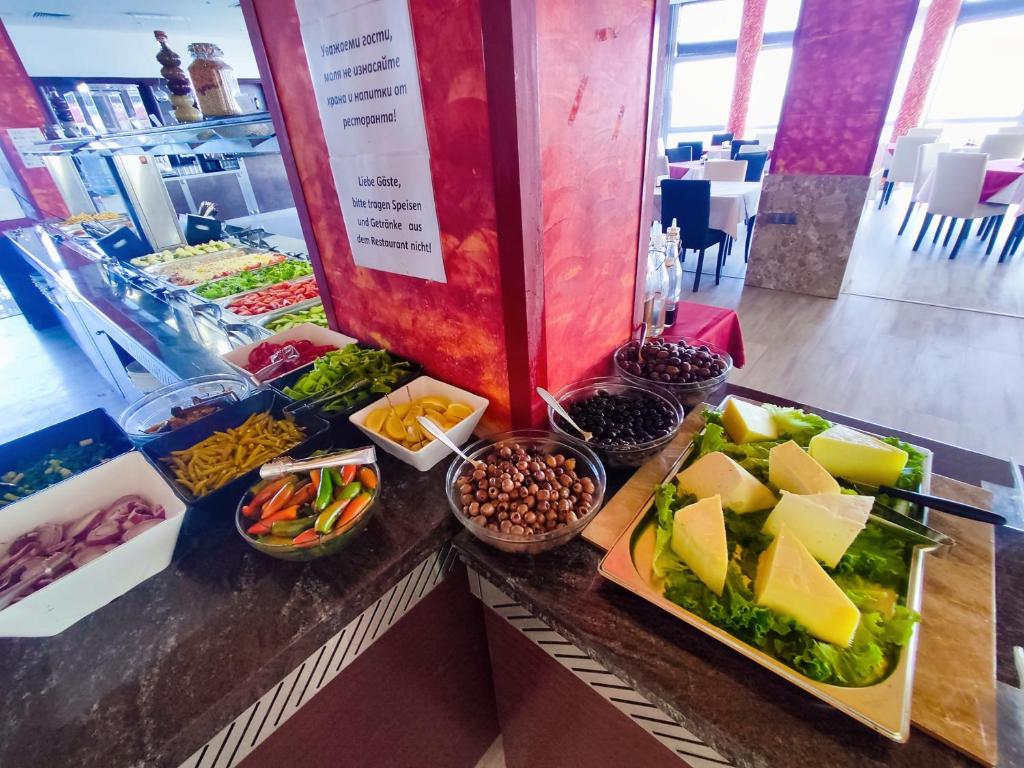 Bufetes galds ar dažādiem dārzeņiem, sieriem un olīvām, dekorēts zaļumos un atrodas restorānā. Fons ir gaišs un moderns, radot patīkamu atmosfēru.