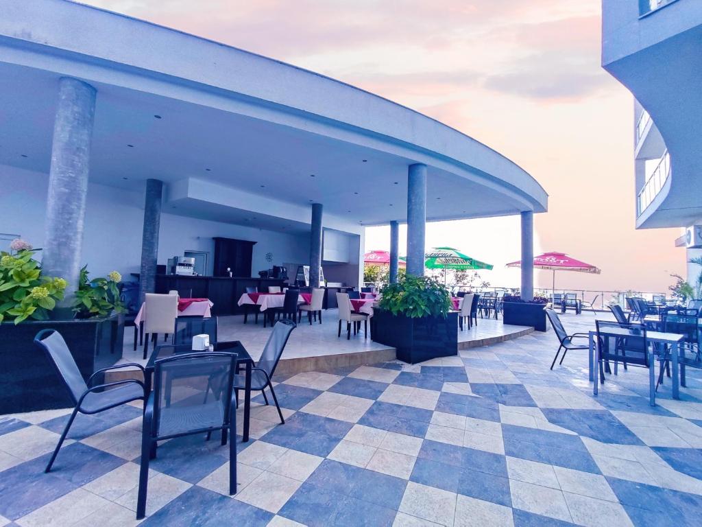Mūsdienīga terase ar ēdamgaldiem un krēsliem, kas atrodas pie viesnīcas restorāna, ar skatu uz jūru un saulrietu. Svaigas zālītes un sauļošanās krēsli papildina relaksējošo atmosfēru.