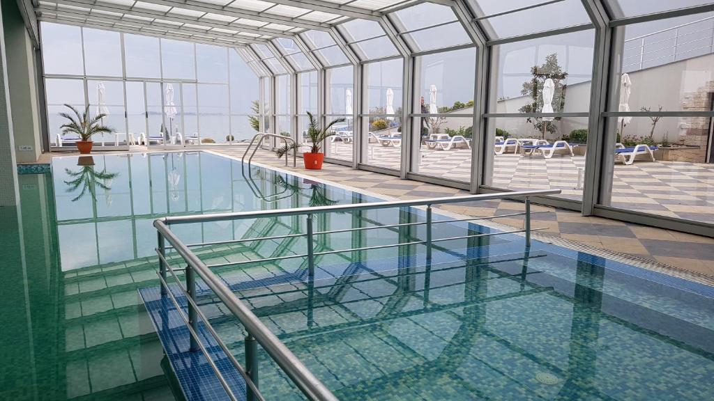 Iekštelpu peldbaseins ar caurspīdīgām sienām un skatu uz ārējo atpūtas zonu, kur redzami sauļošanās krēsli un palmas.