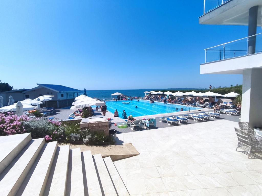 Vasaras atpūtas vieta ar baseinu un jūras skatu, kur cilvēki atpūšas zem saulessargiem. Apkārt ir ziedi un moderni mēbeļu elementi.