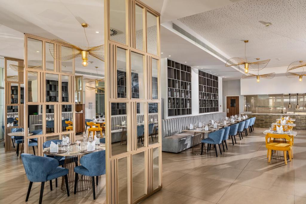 Mūsdienīgs restorāna interjers ar elegantām mēbelēm un apgaismojumu, kas izceļ stilīgu atmosfēru. Sēdvietas ir izvietotas ap apaļiem un garajiem galdiem, radot ērtu un pievilcīgu vidi.