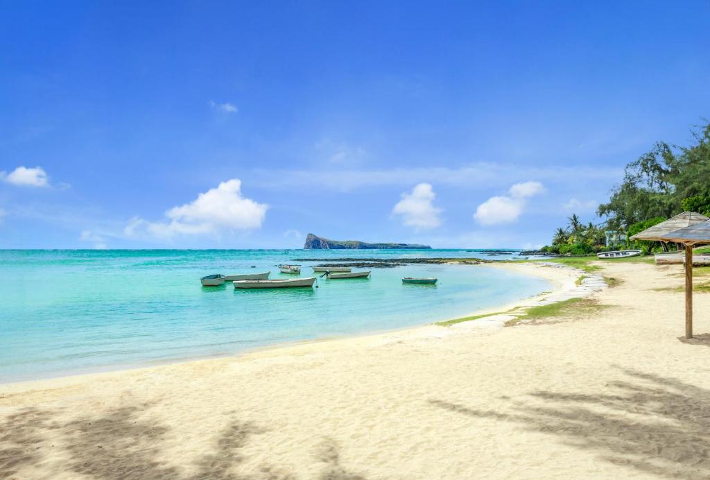 Skaista pludmale ar smilšainu krastu un zilu ūdeni, kur atrodas laivas. Fons ar kokiem un kalniem, kas veido idillisku ainavu.