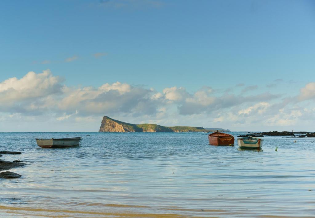 Pludmales ainava ar trim laivām, kas atrodas mierīgā ūdenī, un fona kalnu veidojumu zem zila debesu fona.