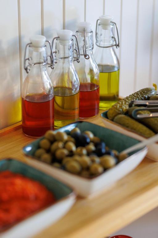 Skaisti stikla pudeļu komplekts ar olīvām, marinētiem gurķiem un sarkanām mērēm, kas izkārtots uz koka plaukta.