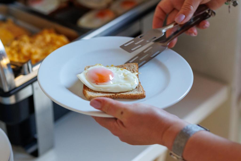 Uz šķīvja ir grauzdiņš ar vārītu olu, ko pasniedz brokastīs. Fons attēlo ēdienu pagatavošanas procesu.