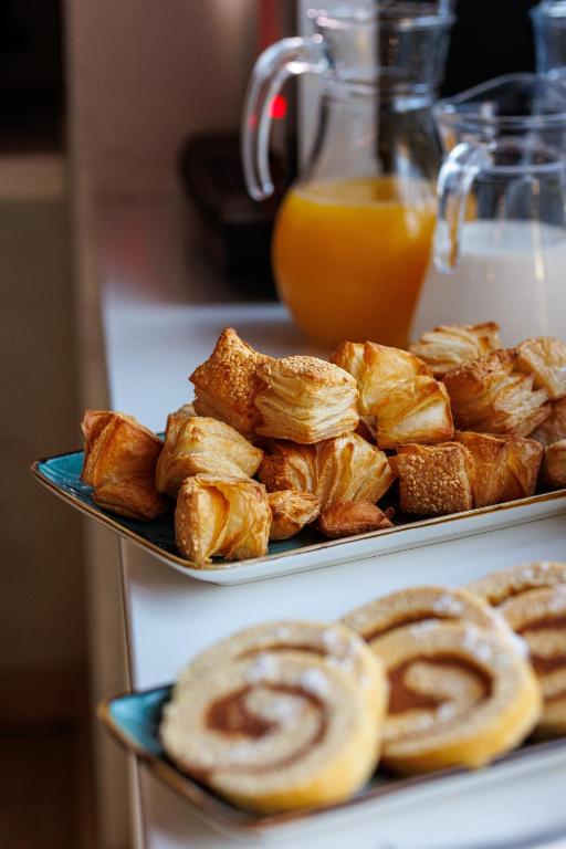 Svaigi cepti konditorejas izstrādājumi uz baltas galda virsmas, iekļaujot maigas, kārtainas maizītes un kanēļa ruletes, blakus dzērienu traukiem ar apelsīnu sulu un pienu.
