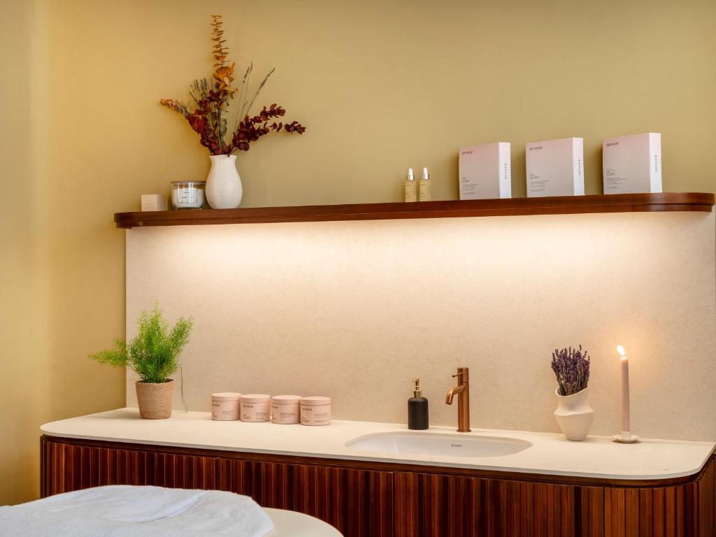 Mūsdienīga un eleganta SPA telpa ar dabīgiem elementiem, piemēram, ziediem un augiem, kas rada relaksējošu atmosfēru. Uz plaukta redzami skaistumkopšanas produkti un sveces, kas papildina kopējo estētiku.