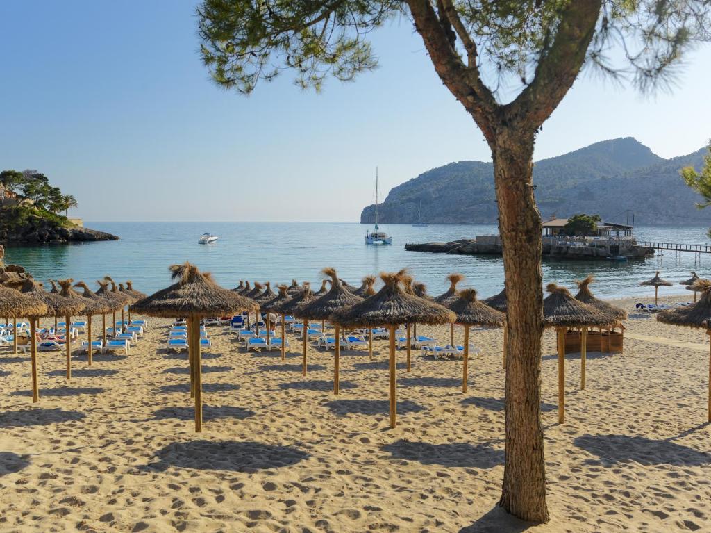 Pludmales ainava ar salmu saulessargiem un zilo jūru, kas atspoguļo kalnu kontūras. Rāmā atmosfērā redzami laivas un pludmales krēsli, radot relaksējošu vidi.