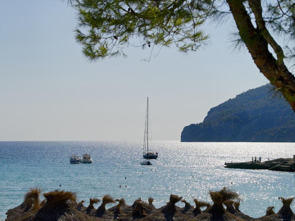 Skaists pludmales skats ar jachtu un laivu, kas pelda ūdenī, fona kalnu un saulainu debesi. Pludmalē redzamas salmu nojumes, kas piešķir tropisku noskaņu.