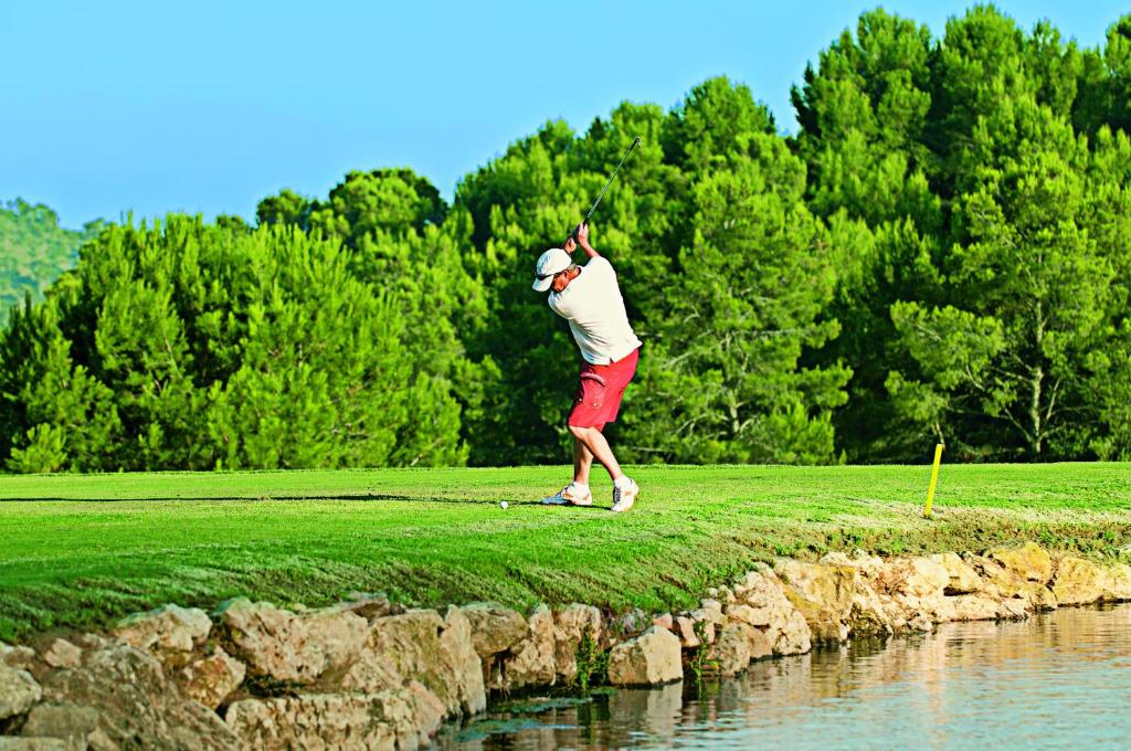 Golfista sitiens bumbiņu uz golfa laukuma, ar zaļām kokiem fona un ūdens malas. Ideāla ainava golfa spēlei.