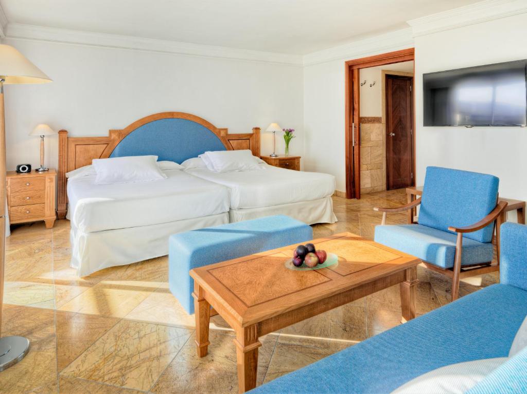 Mūsdienīga viesnīcas numura iekārtojums ar divām gultām, koka mēbelēm un zilu tekstilu. Numurā ir arī televizors un stūra galds ar augļiem.