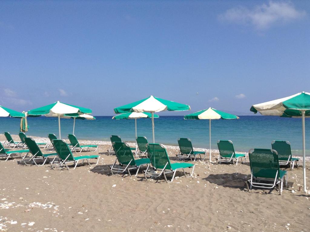 Pludmales ainava ar zaļām sauļošanās krēslām un baltām lietussargiem, kas stāv uz smiltīm pie mierīga jūras krasta. Skats uz zilām debesīm un jūru rada relaksējošu atmosfēru.