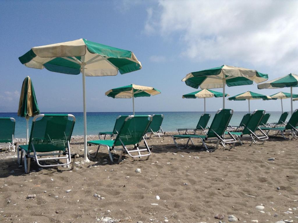 Pludmales ainava ar zaļām sauļošanās krēsliem un lietussargiem, kas izkārtoti smiltīs pie jūras. Skats uz saulainu debesu un mierīgu ūdeni.