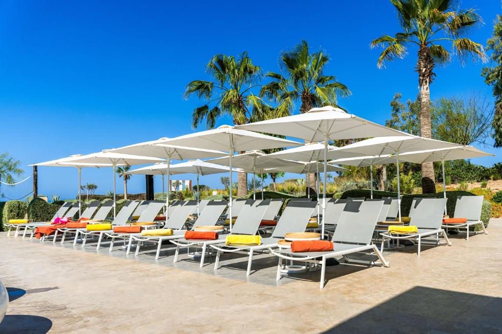 Saulēti pludmales krēsli ar saulessargiem, novietoti pie baseina vai pludmales, baudot siltu laiku un brīvdienu atmosfēru. Pilsētiņā redzami palmas un zils debesis.