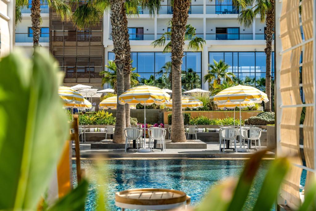 Peldbaseins ar saules krēsliem un dzeltenām lietussargiem, ieskauts palmām un zaļumiem, radot atpūtas un relaksācijas atmosfēru. Modernas ēkas fons piešķir attēlam eleganci.