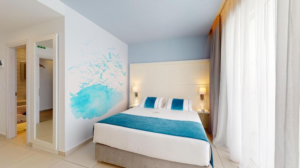 Mūsdienīgs guļamistabas interjers ar baltām sienām un gaisīgu zilu akcentu. Uz sienas ir mākslinieciska zila krāsa, kas atgādina ūdeni, un telpā ir lieli logi ar viegliem aizkariem.