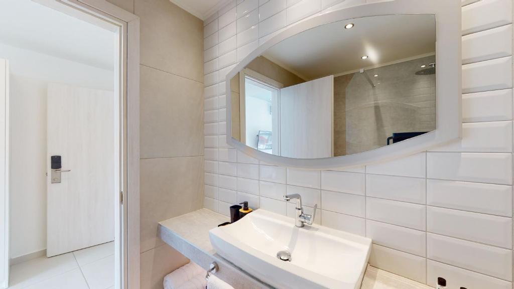 Mūsdienīgs vannas istabas interjers ar gaišu flīžu apdari un ovālu spoguli virs izlietnes. Redzama arī durvju un skapja daļa.