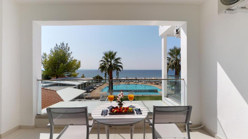 Skats no viesnīcas balkona uz jūru, kur redzams baseins un palmām apkārt. Uz galda atrodas augļi un dzērieni, radot relaksējošu atmosfēru.