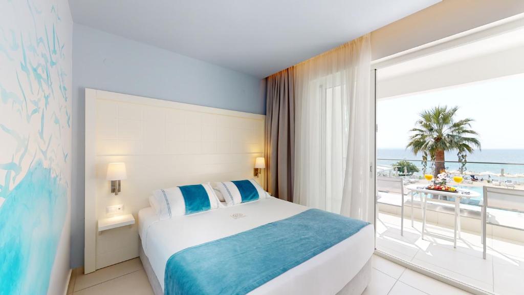 Mūsdienīga viesnīcas istaba ar skatu uz jūru, gaišām krāsām un ērtu gultu. Logā redzams palmām rotāts pludmales skats.