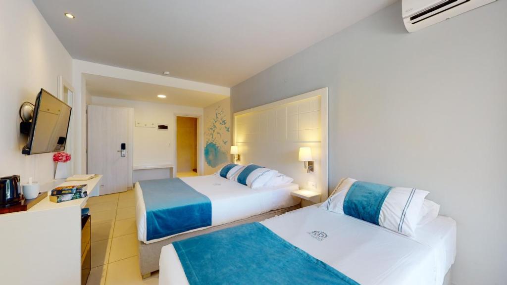 Mūsdienīgs viesnīcas numurs ar divām gultām un gaišu interjeru, kurā ir televizors un gaismas elementi. Sienā ir dekoratīvs mākslas darbs, piešķirot telpai patīkamu atmosfēru.